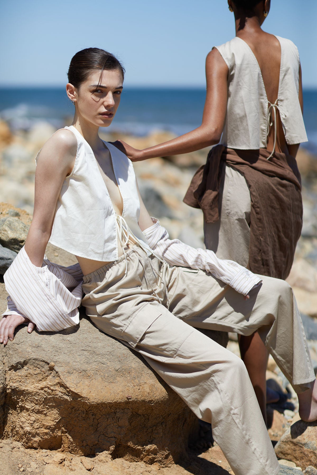Two models in light, minimalistic outfits sitting on rocks by the ocean. Leset collection.