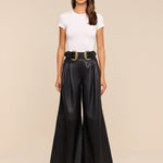 A woman stands facing forward against a plain beige background, wearing sunglasses, a white fitted t-shirt, and the Chelsy Leather Wide-Leg Pant in black styled with a black belt featuring two large gold buckles.