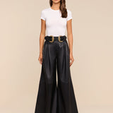 A woman stands facing forward against a plain beige background, wearing sunglasses, a white fitted t-shirt, and the Chelsy Leather Wide-Leg Pant in black styled with a black belt featuring two large gold buckles.