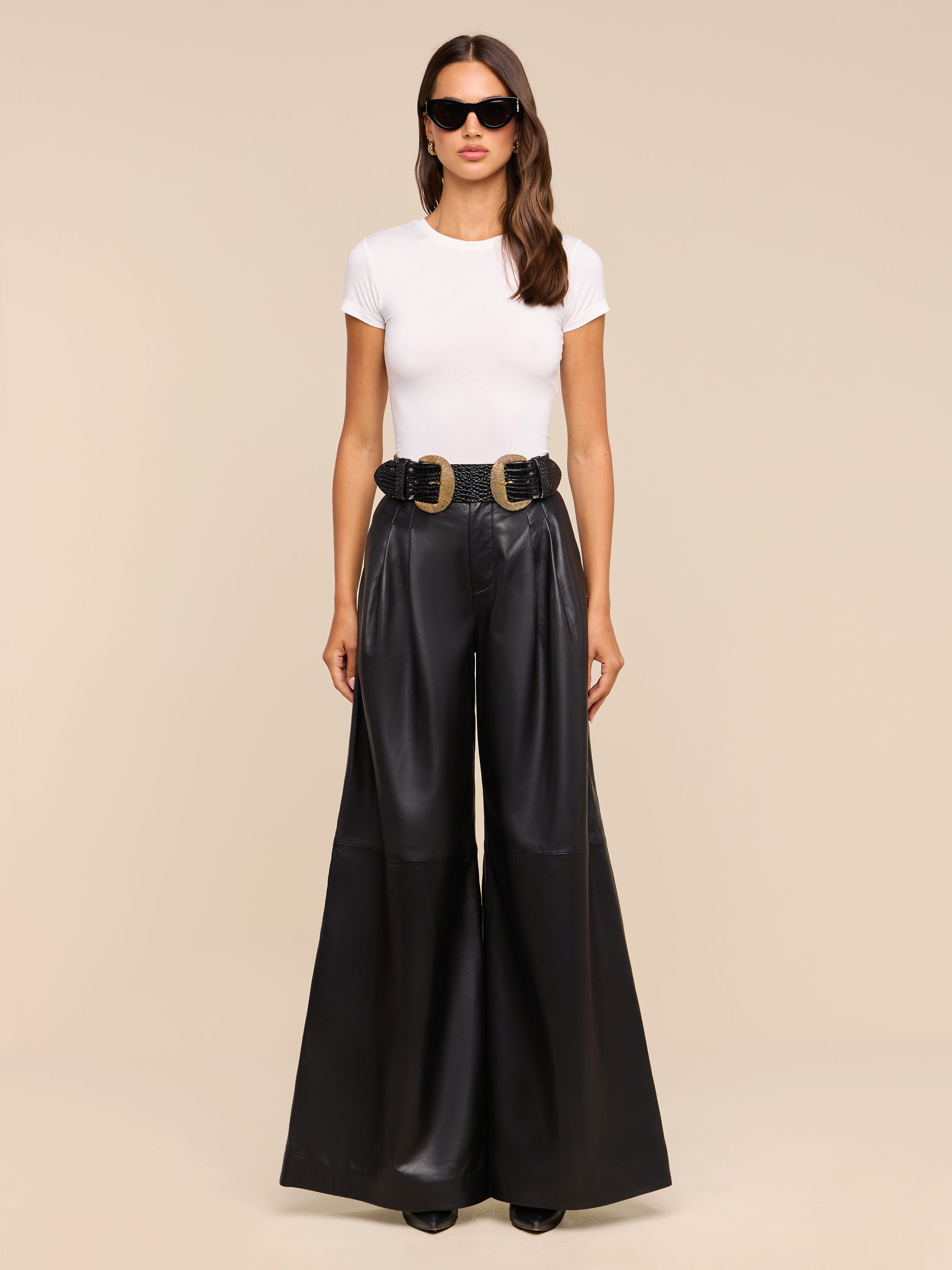 A woman stands facing forward against a plain beige background, wearing sunglasses, a white fitted t-shirt, and the Chelsy Leather Wide-Leg Pant in black styled with a black belt featuring two large gold buckles.