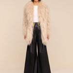A woman stands against a light background wearing a fluffy beige faux fur coat, white top, Chelsy Leather Wide-Leg Pant, black belt with a gold buckle, black shoes, and black sunglasses.