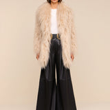 A woman stands against a light background wearing a fluffy beige faux fur coat, white top, Chelsy Leather Wide-Leg Pant, black belt with a gold buckle, black shoes, and black sunglasses.