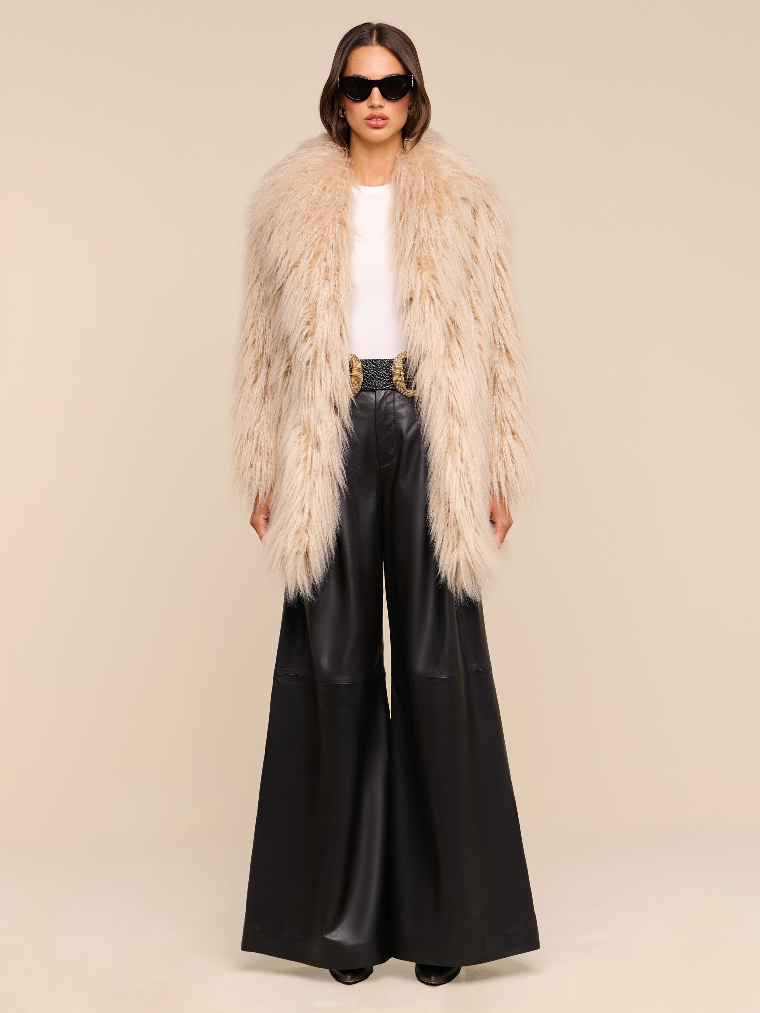 A woman stands against a light background wearing a fluffy beige faux fur coat, white top, Chelsy Leather Wide-Leg Pant, black belt with a gold buckle, black shoes, and black sunglasses.