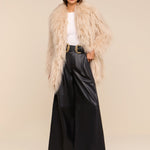 A woman wearing a beige faux fur coat, white top, the Chelsy Leather Wide-Leg Pant in black, a black belt with a gold buckle, and sunglasses stands confidently against a plain beige background.