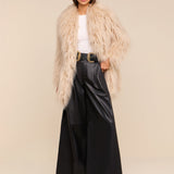A woman wearing a beige faux fur coat, white top, the Chelsy Leather Wide-Leg Pant in black, a black belt with a gold buckle, and sunglasses stands confidently against a plain beige background.