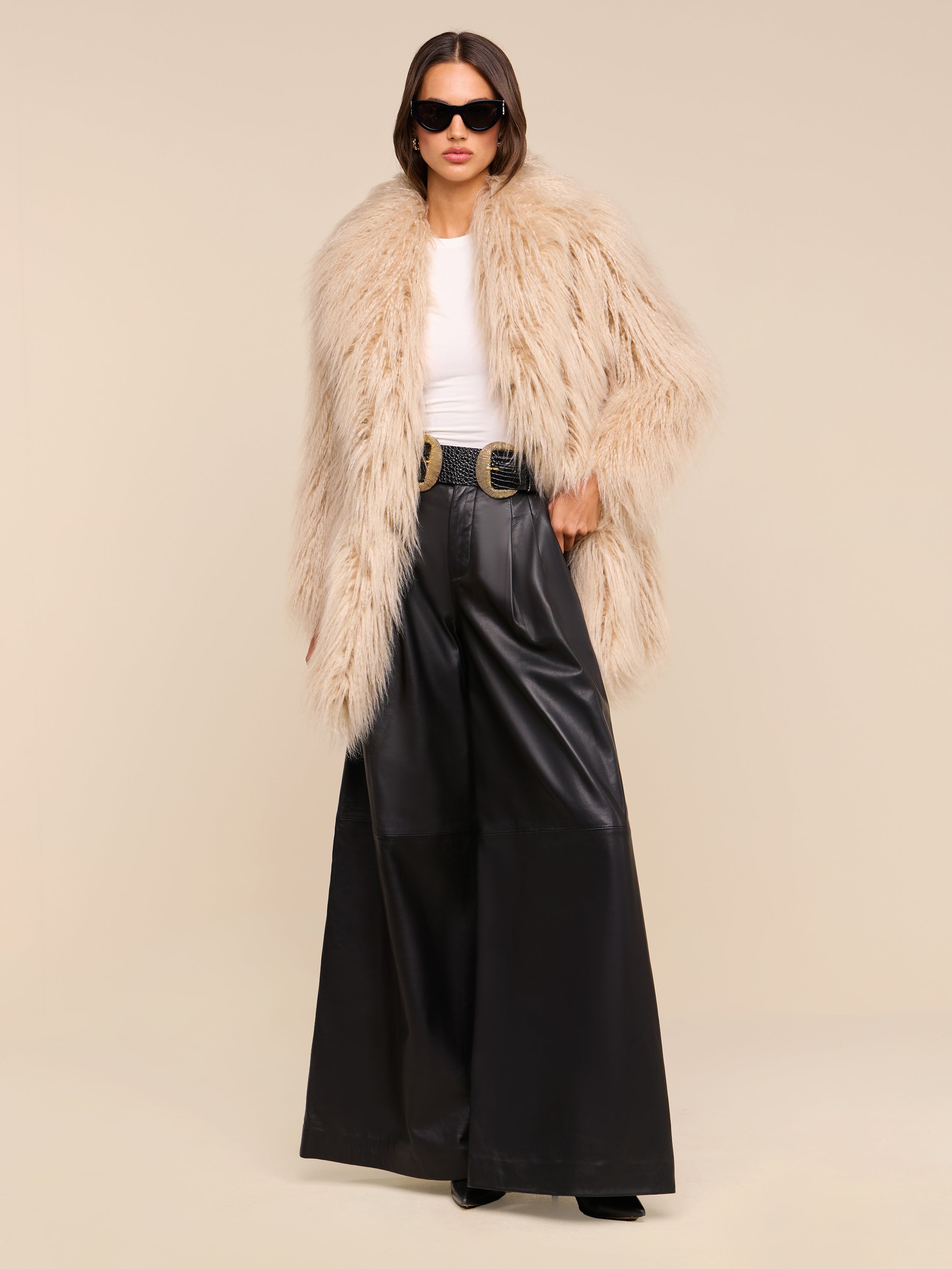 A woman wearing a beige faux fur coat, white top, the Chelsy Leather Wide-Leg Pant in black, a black belt with a gold buckle, and sunglasses stands confidently against a plain beige background.