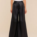 A person stands against a beige background, wearing a white sleeveless top and the Chelsy Leather Wide-Leg Pant—high-rise, lightweight leather pants featuring a wide leg. Only their body from the shoulders down is visible.