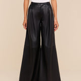 A person stands against a beige background, wearing a white sleeveless top and the Chelsy Leather Wide-Leg Pant—high-rise, lightweight leather pants featuring a wide leg. Only their body from the shoulders down is visible.