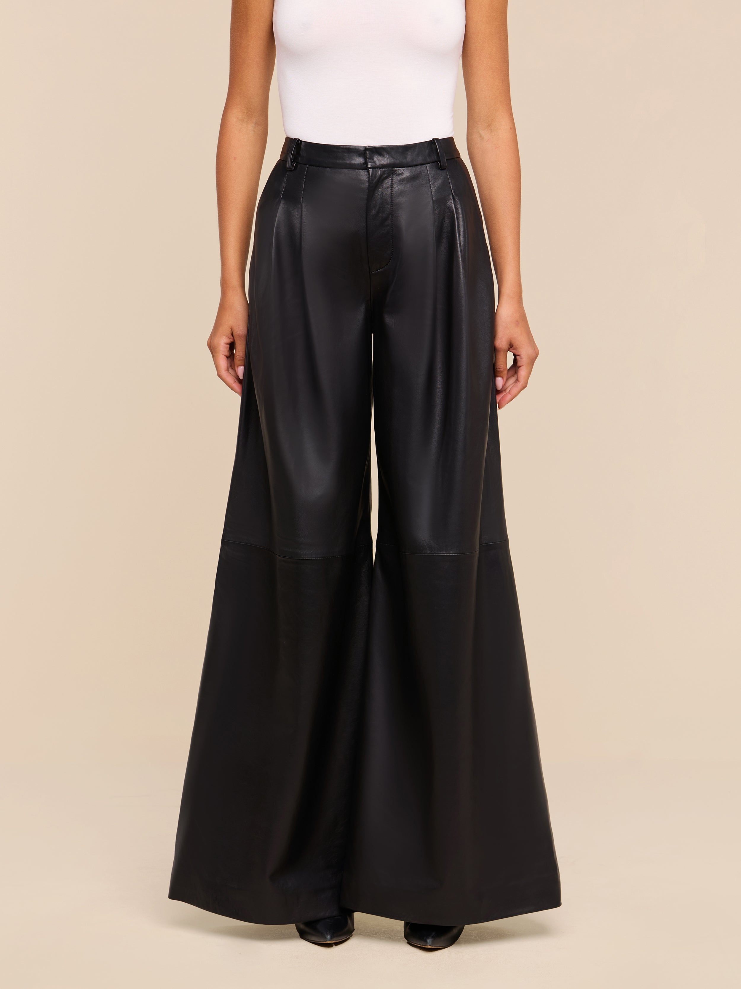 A person stands against a beige background, wearing a white sleeveless top and the Chelsy Leather Wide-Leg Pant—high-rise, lightweight leather pants featuring a wide leg. Only their body from the shoulders down is visible.