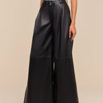 A person stands against a beige background wearing the Chelsy Leather Wide-Leg Pant in black—high rise, wide leg, and lightweight with side pockets—paired with a white sleeveless top and black pointed-toe shoes.