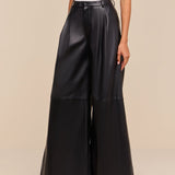 A person stands against a beige background wearing the Chelsy Leather Wide-Leg Pant in black—high rise, wide leg, and lightweight with side pockets—paired with a white sleeveless top and black pointed-toe shoes.