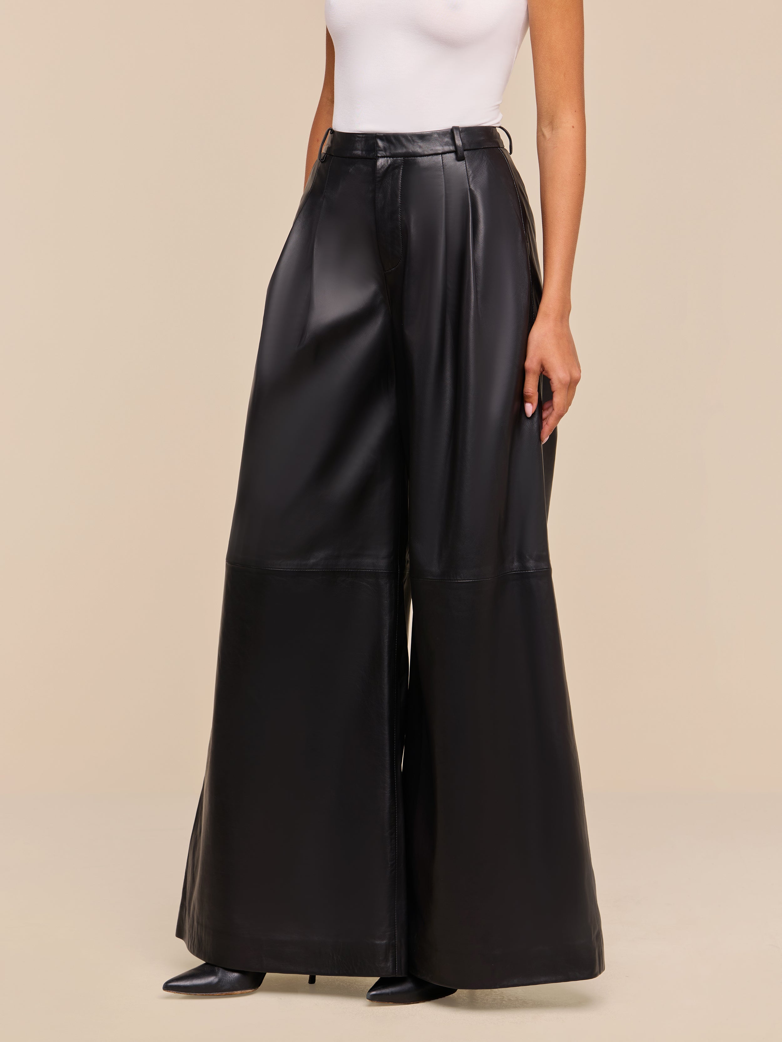 A person stands against a beige background wearing the Chelsy Leather Wide-Leg Pant in black—high rise, wide leg, and lightweight with side pockets—paired with a white sleeveless top and black pointed-toe shoes.