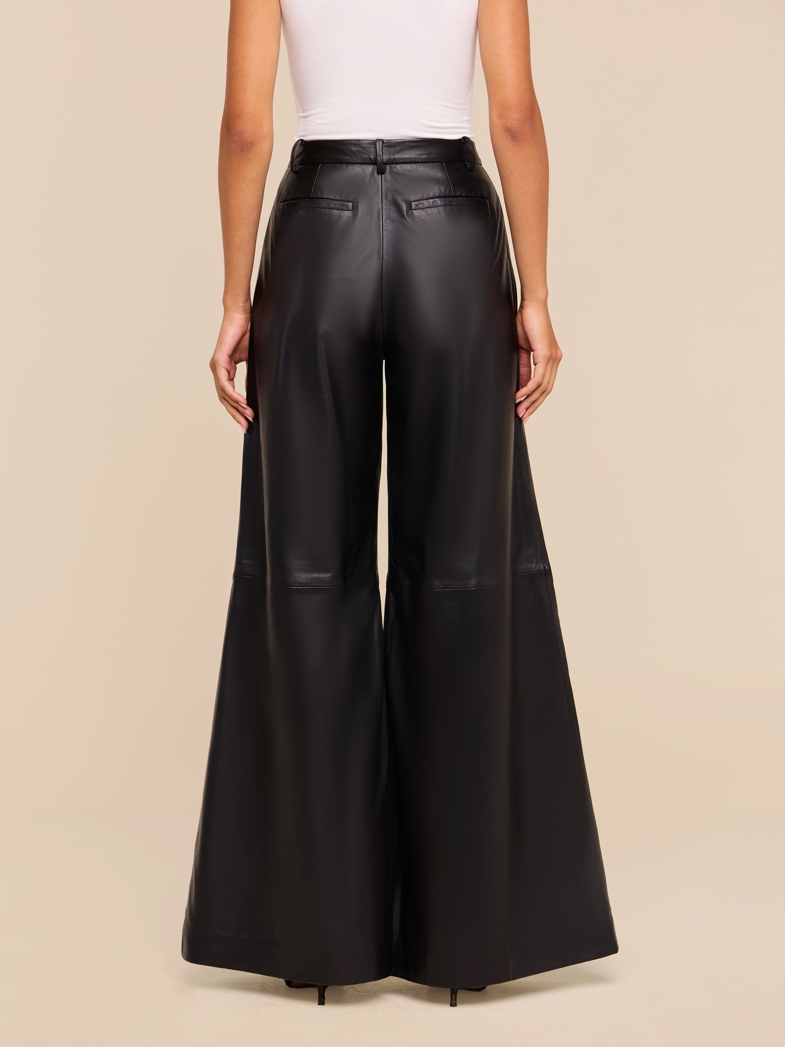 Wearing the Chelsy Leather Wide-Leg Pant in a high-rise, black lightweight leather, paired with a white sleeveless top, a person is pictured from the back against a plain beige background.