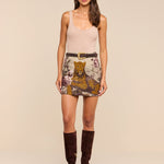 A woman stands against a beige background wearing a nude tank top, the Marseille Mini Skirt—a vintage-inspired mini skirt featuring leopard and floral prints—a brown belt, and knee-high brown suede boots.
