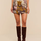 Wearing the Marseille Mini Skirt—vintage-inspired with a bold leopard and floral design—cinched at the waist with a brown belt and paired with knee-high brown suede boots, they stand against a neutral background.