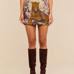 A woman wearing the Marseille Mini Skirt, which features a vintage-inspired large leopard and floral pattern, paired with a light pink top and dark brown knee-high suede boots.