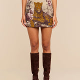 A woman wearing the Marseille Mini Skirt, which features a vintage-inspired large leopard and floral pattern, paired with a light pink top and dark brown knee-high suede boots.