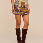 A woman wearing a beige top, the Marseille Mini Skirt—a vintage-inspired leopard tapestry mini skirt with a floral design—a black belt, and knee-high dark brown suede boots, standing against a plain beige background.