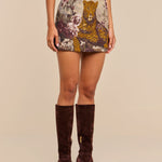 A woman wearing a beige top, the Marseille Mini Skirt featuring a vintage-inspired leopard and floral print, and knee-high brown suede boots stands against a plain background.