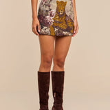 A woman wearing a beige top, the Marseille Mini Skirt featuring a vintage-inspired leopard and floral print, and knee-high brown suede boots stands against a plain background.