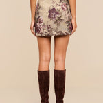 A woman wears the Marseille Mini Skirt, a vintage-inspired mini skirt featuring a floral pattern, paired with knee-high brown suede boots; she is shown from the back against a plain beige background.