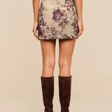 A woman wears the Marseille Mini Skirt, a vintage-inspired mini skirt featuring a floral pattern, paired with knee-high brown suede boots; she is shown from the back against a plain beige background.