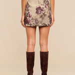 A woman stands facing away, wearing the Marseille Mini Skirt—a vintage-inspired mini skirt with a floral pattern—paired with a beige top and knee-high brown suede boots. The background is plain beige.