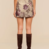 A woman stands facing away, wearing the Marseille Mini Skirt—a vintage-inspired mini skirt with a floral pattern—paired with a beige top and knee-high brown suede boots. The background is plain beige.
