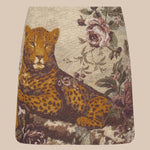 The Marseille Mini Skirt is a vintage-inspired mini skirt crafted from luxurious Italian jacquard, featuring a reclining leopard surrounded by pink and burgundy flowers on a beige background.