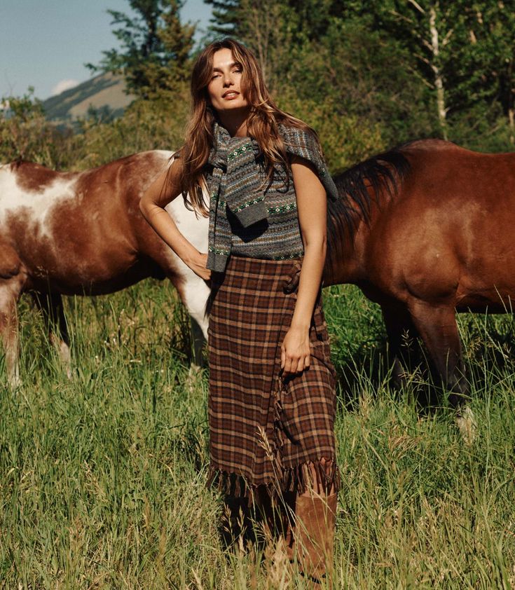 Woman standing in a field with horses wearing a plaid skirt and patterned top.