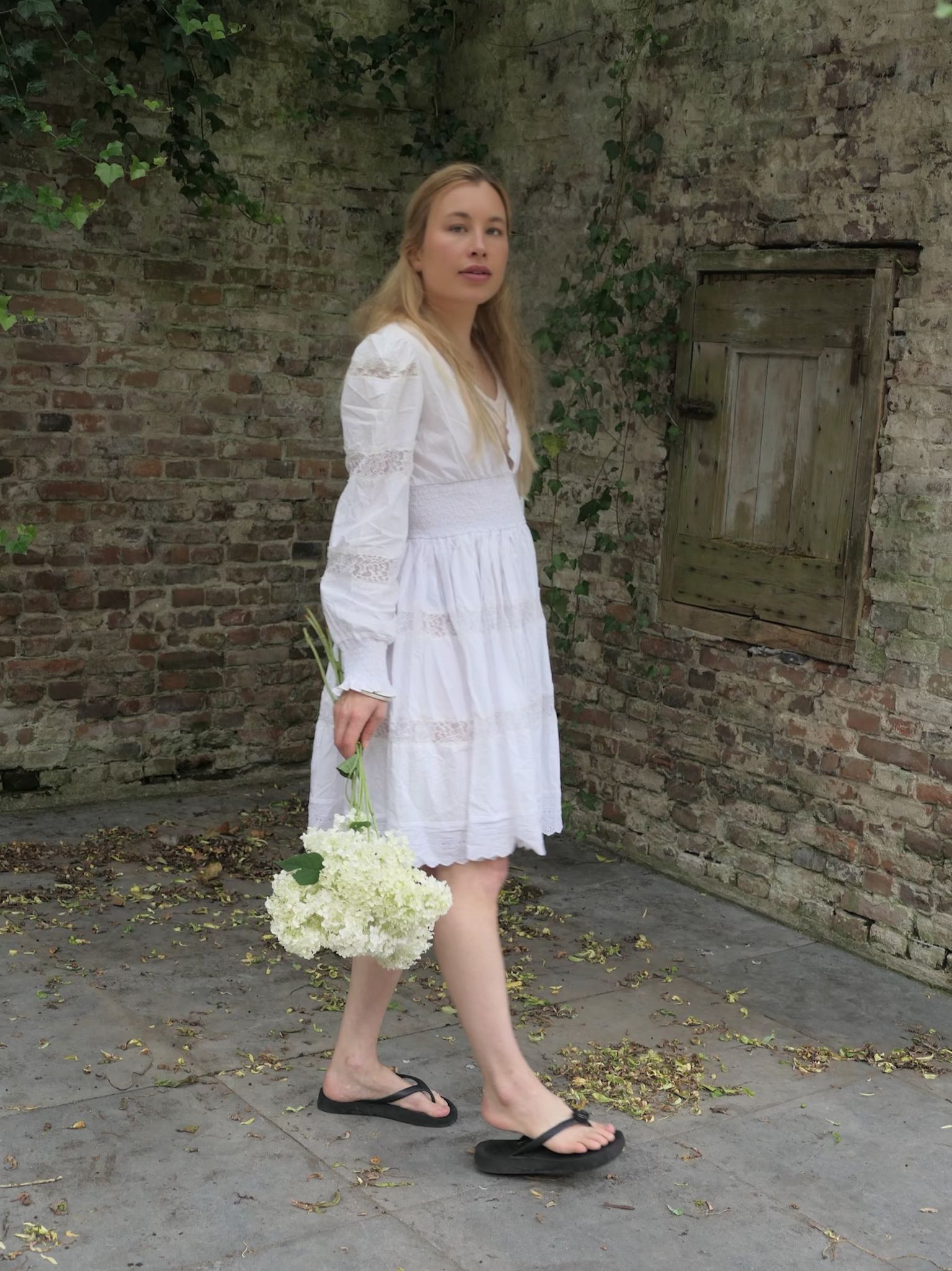 Woman in a white dress holding flowers against a brick wall. Late summer fall capsule wardrobe. 
