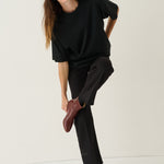 A woman modeling a black oversized short-sleeve top and black wide-leg trousers, paired with dark brown leather ankle boots, against a plain off-white studio background.