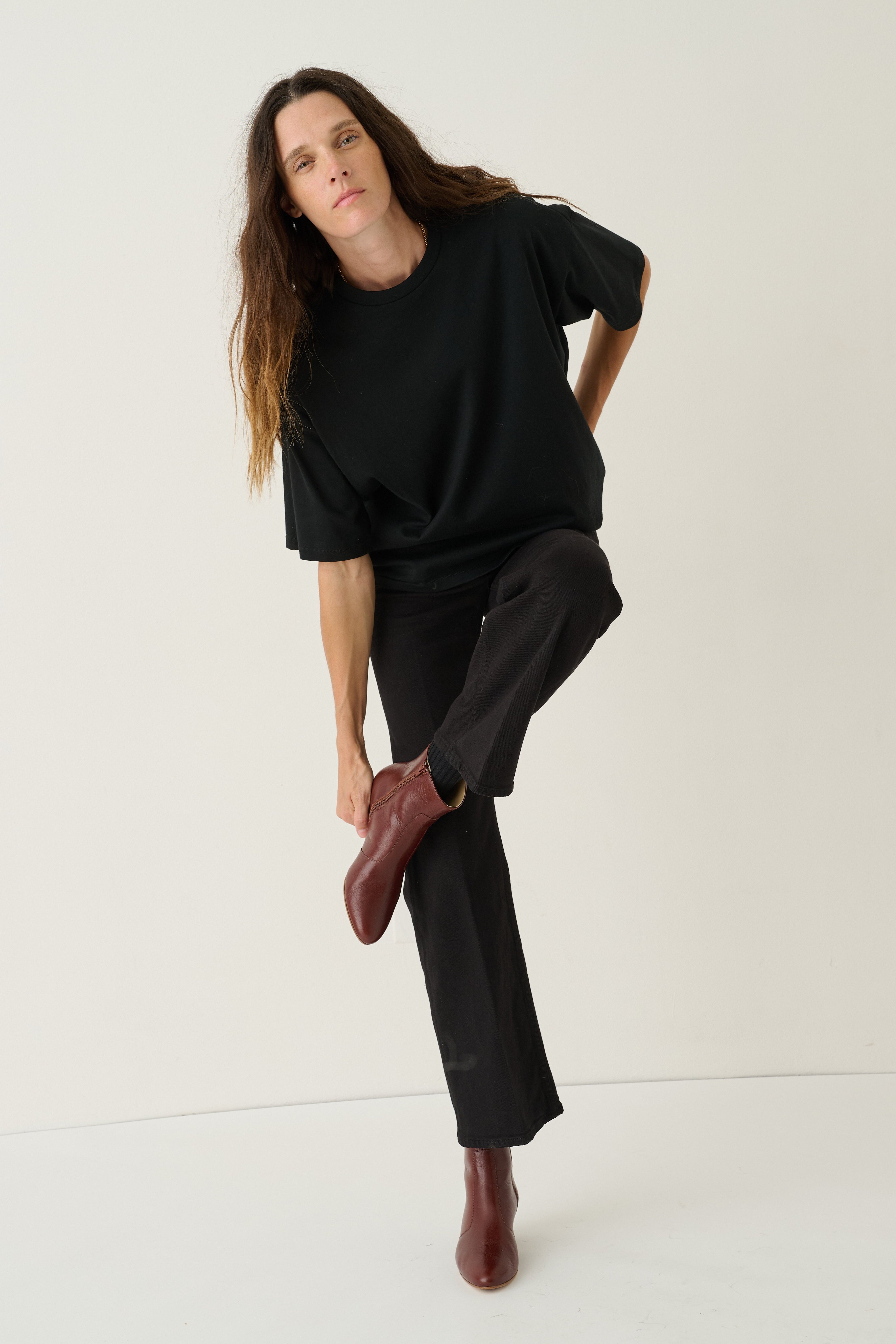 A woman modeling a black oversized short-sleeve top and black wide-leg trousers, paired with dark brown leather ankle boots, against a plain off-white studio background.