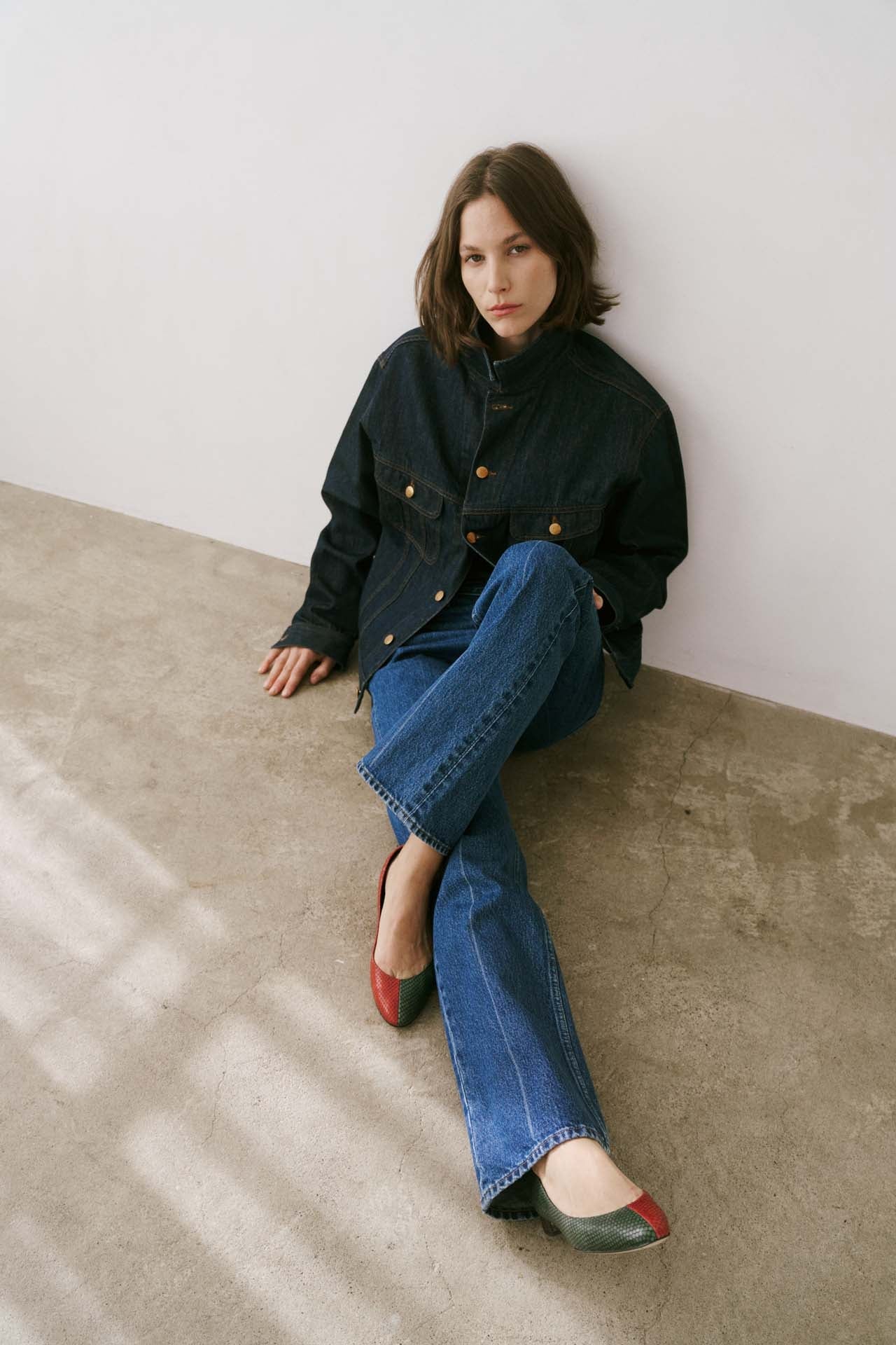 Woman sitting on a concrete floor wearing a dark jacket and blue jeans.
