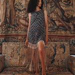 A woman stands barefoot on an ornate tapestry sofa wearing a sleeveless dark dress with a small floral print and long fringe along the hem, posed in front of a large matching tapestry wall hanging.