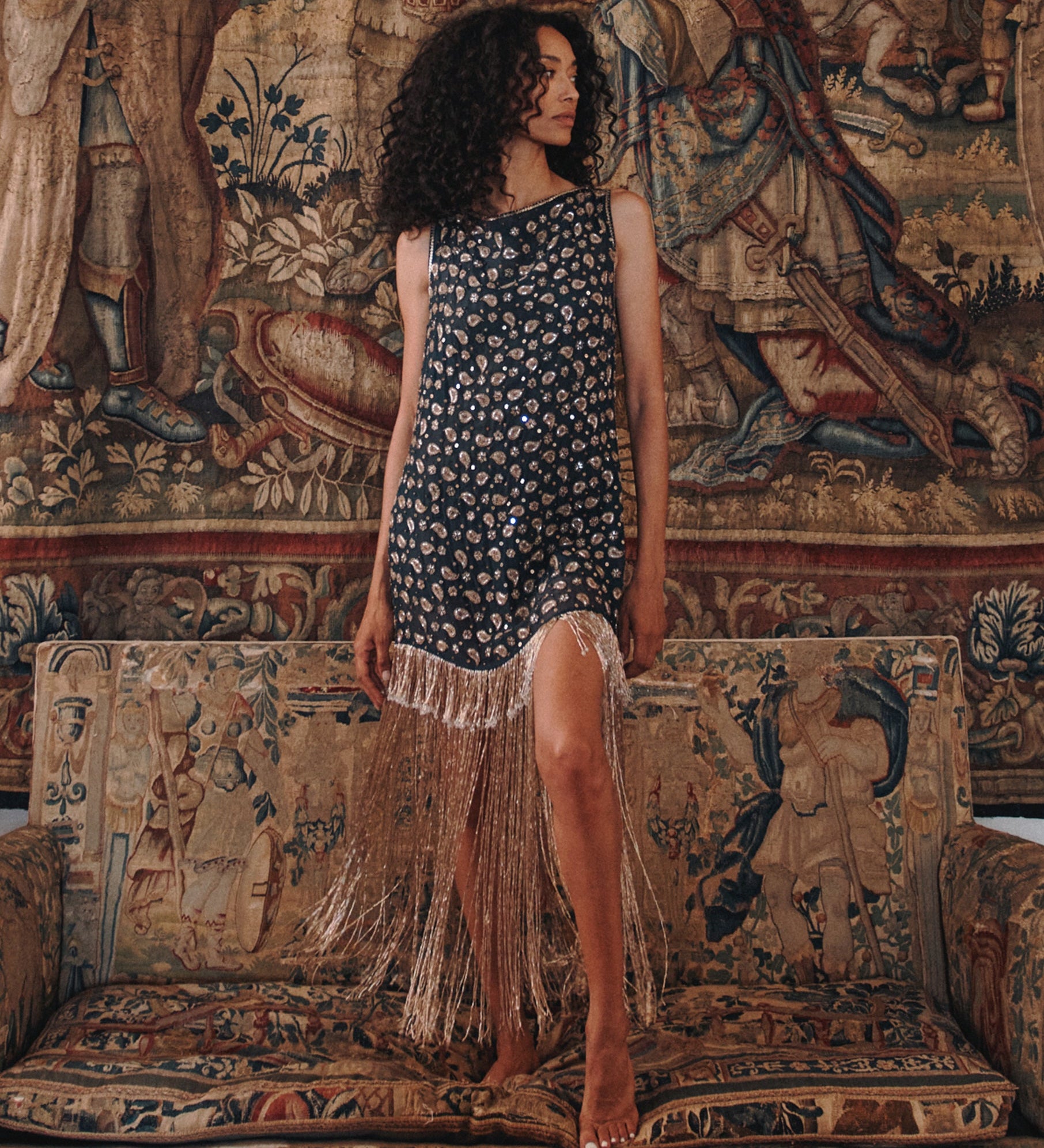 A woman stands barefoot on an ornate tapestry sofa wearing a sleeveless dark dress with a small floral print and long fringe along the hem, posed in front of a large matching tapestry wall hanging.