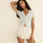Woman wearing a light blue-colored patterned top and white lace shorts against a beige background