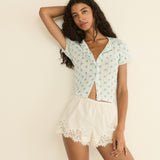 Woman wearing a light blue-colored patterned top and white lace shorts against a beige background