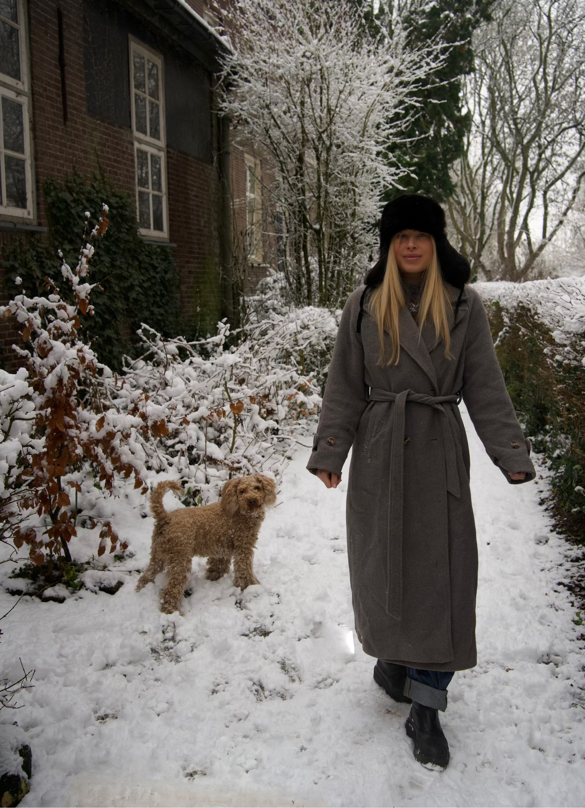 Woman in a long coat walking a dog in a snowy garden.