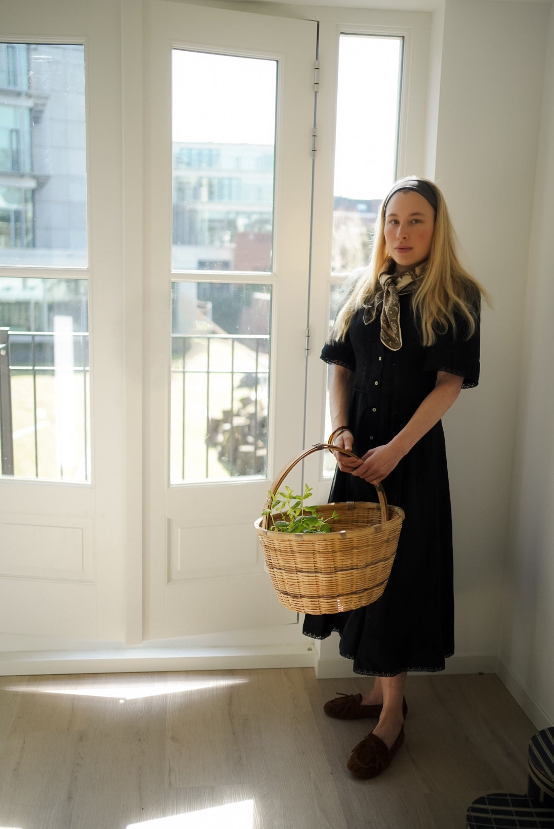 Woman holding a woven basket with plants indoors near a large window.