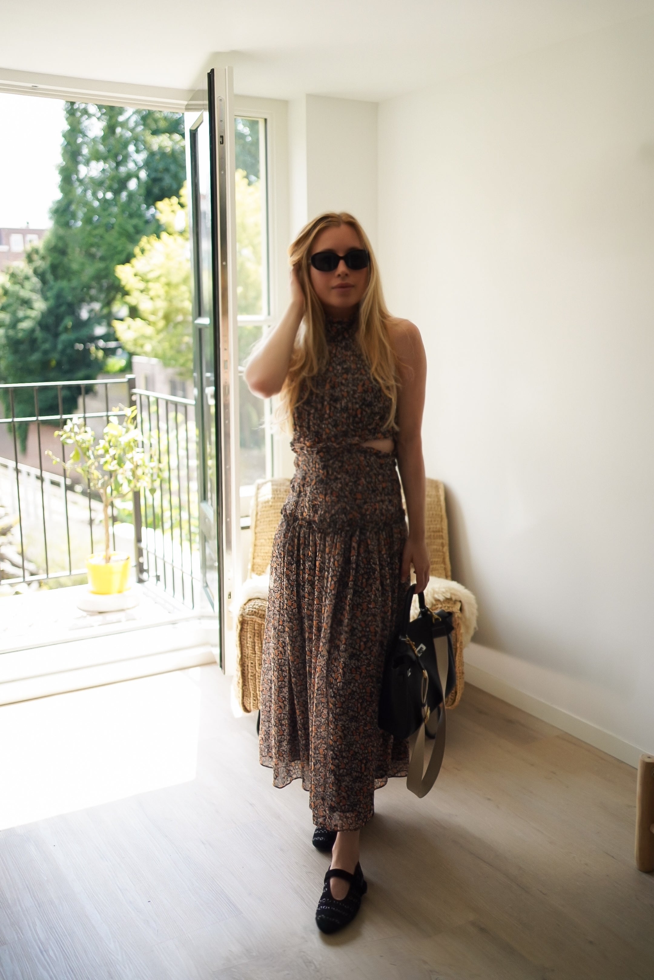 A woman stands in a bright room wearing a sleeveless brown patterned midi dress with a waist tie, black sunglasses, and black Mary Jane shoes, holding a black handbag.