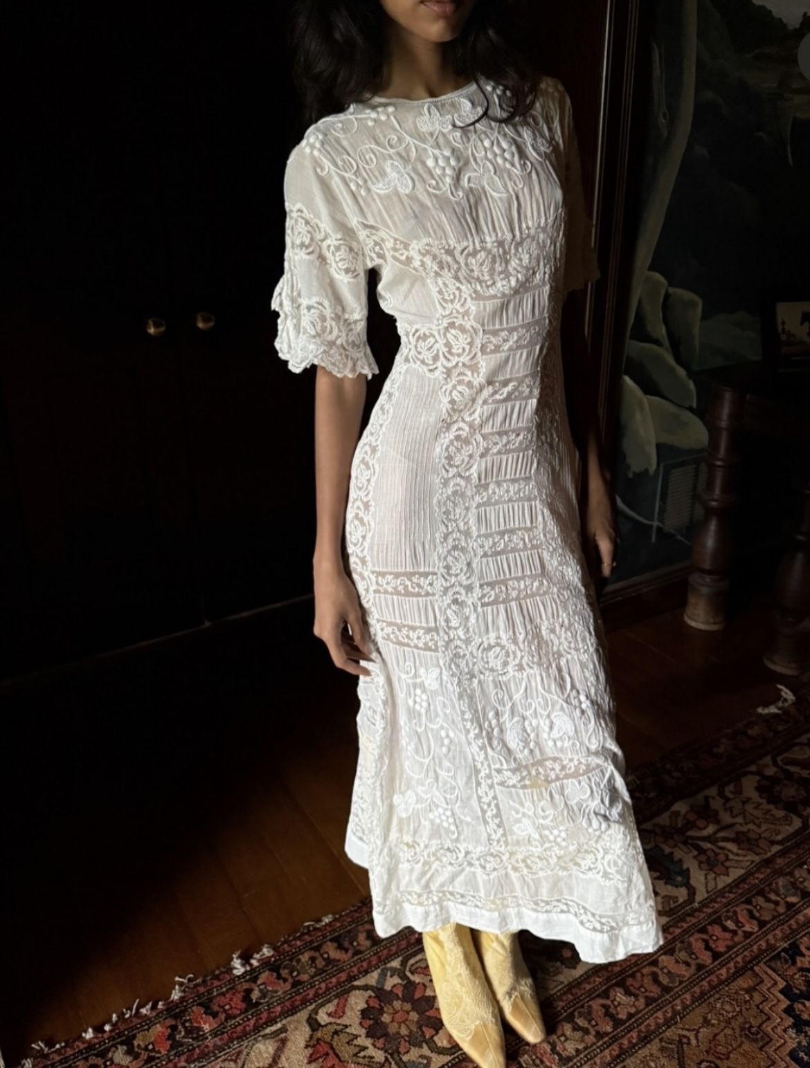 A woman wearing a white lace midi dress with short puff sleeves and tan boots, standing in a dimly lit room with a patterned rug and dark wooden furniture.
