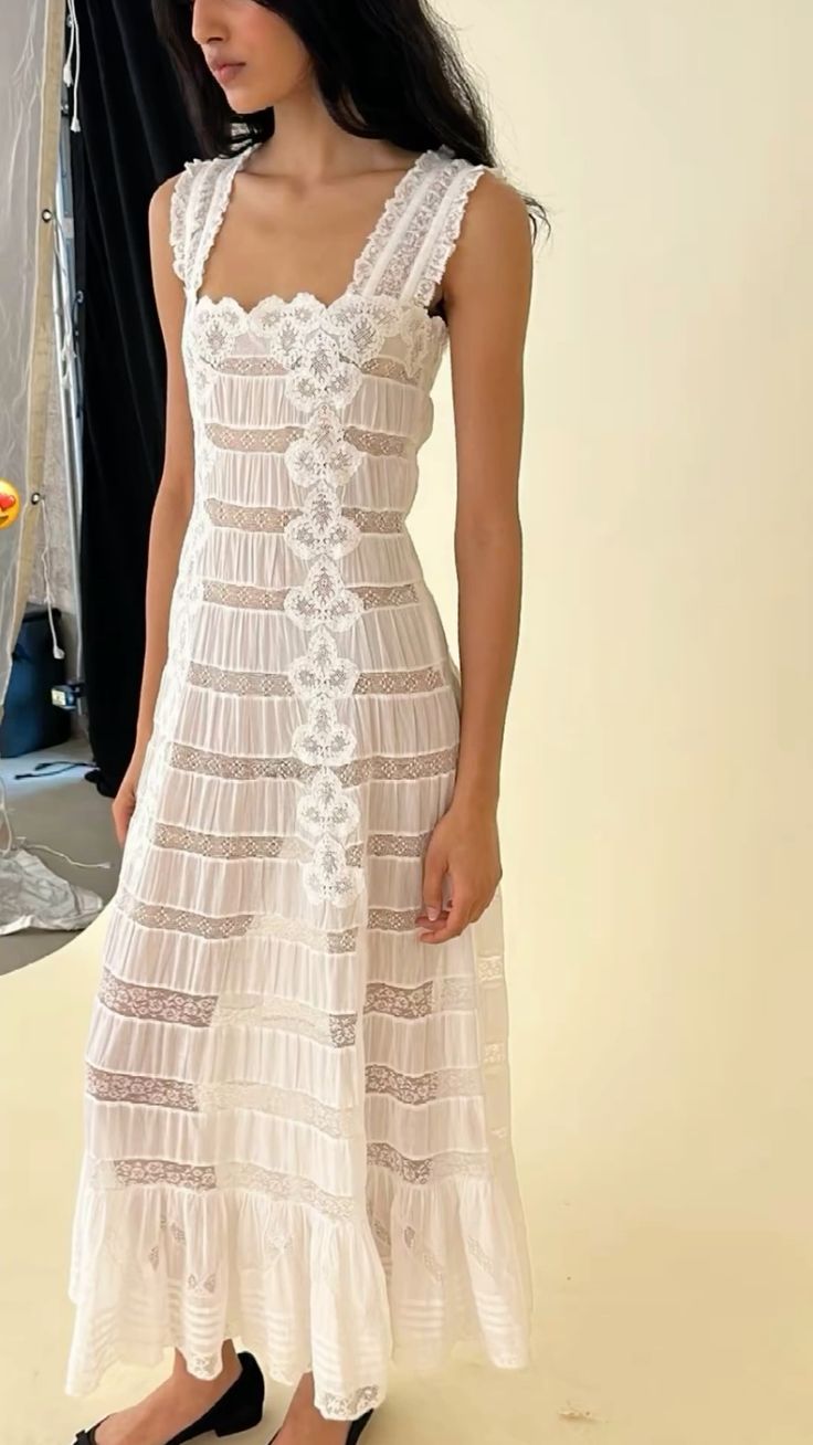 A woman wearing a sleeveless white lace maxi dress with a tiered skirt and floral lace detailing, standing against a plain cream-colored background.