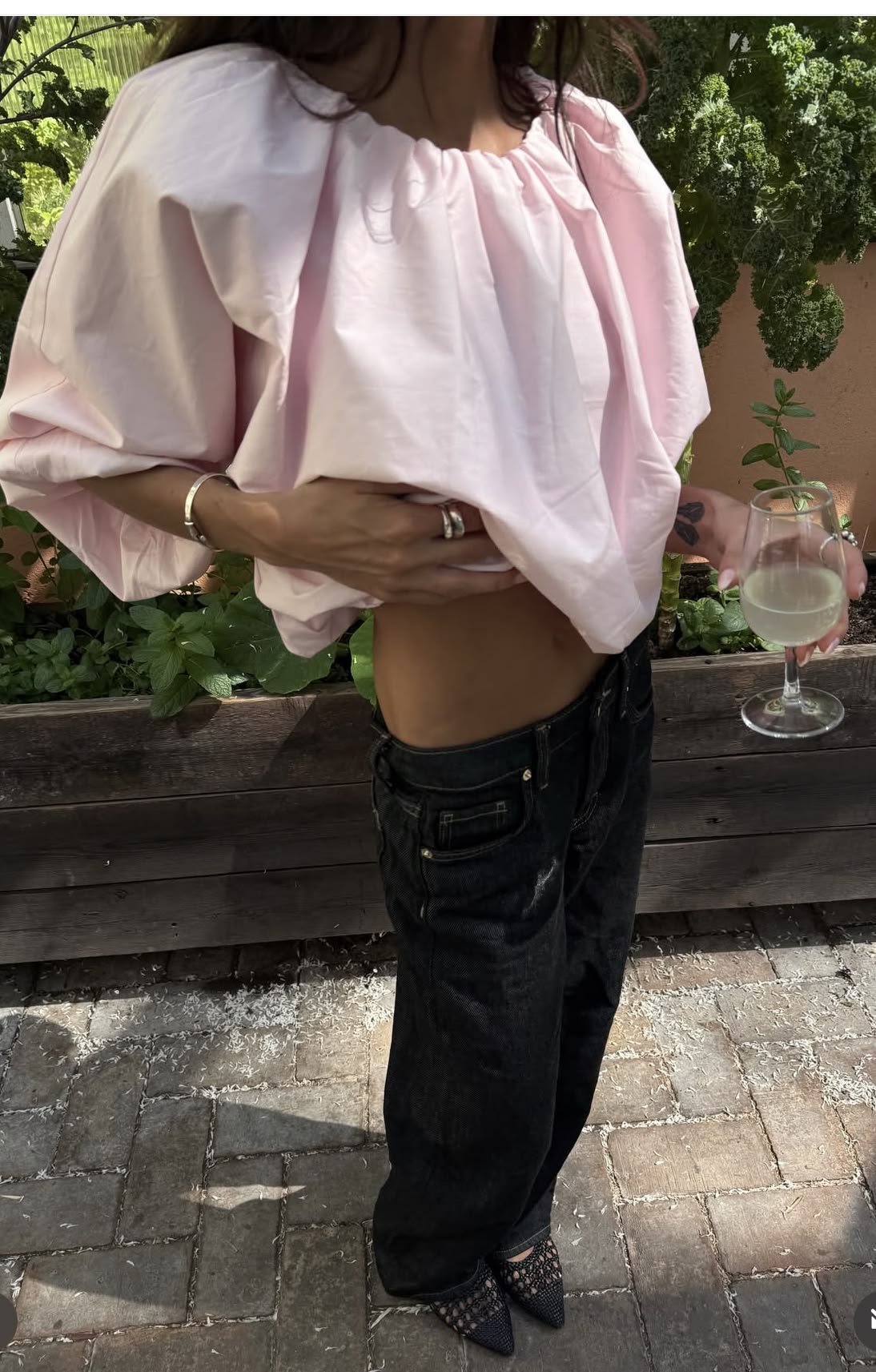 A person outdoors wearing a light pink oversized bubble-sleeve top and loose black jeans, holding a glass of white wine. The background shows a wooden planter with green plants and stone pavers.