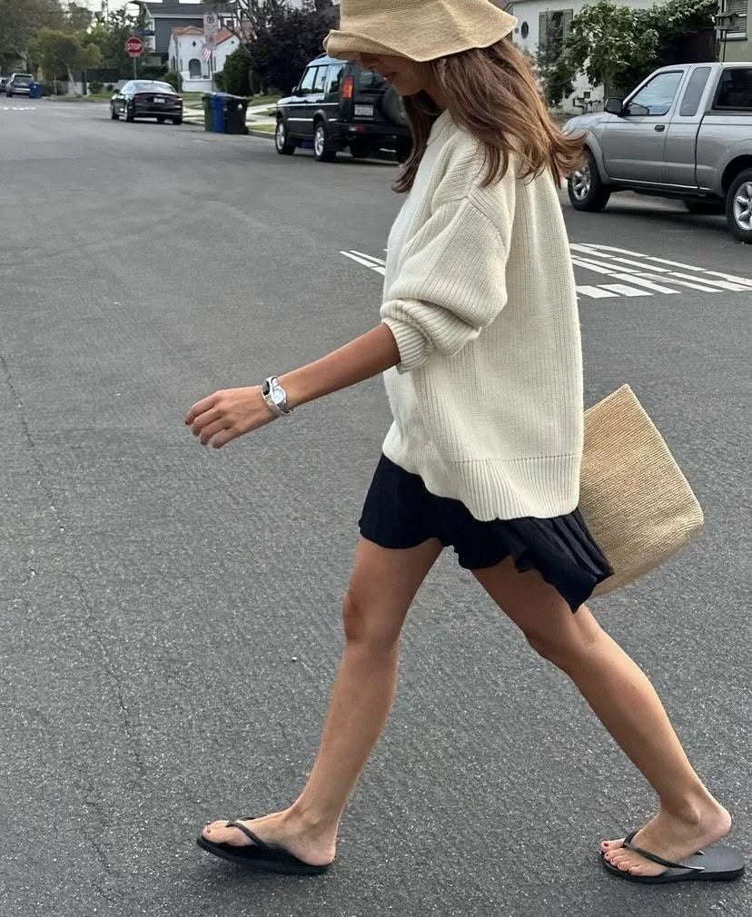 Woman walking on a street wearing a beige sweater and black shorts, holding a beige bag.