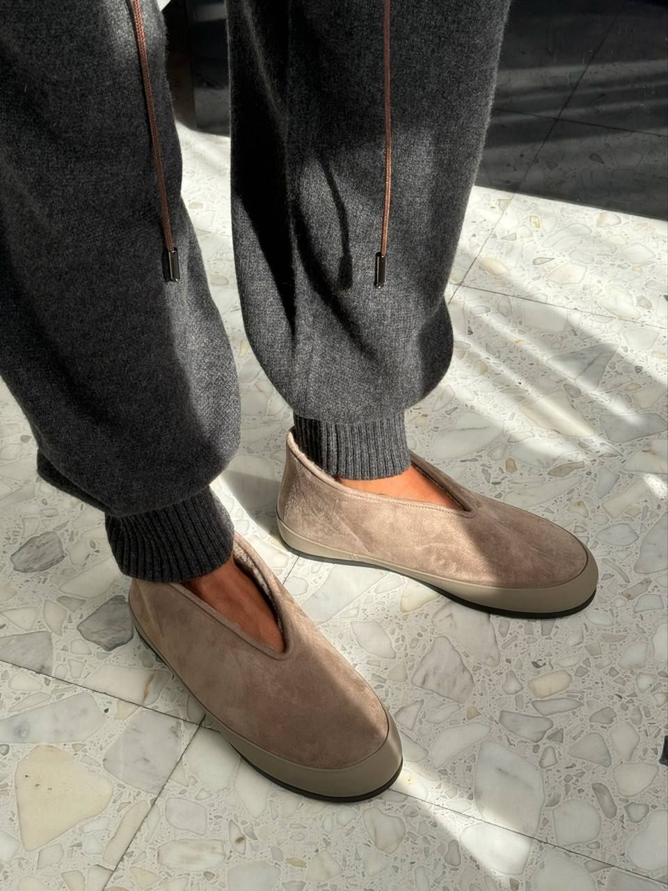 A person wearing dark gray knit jogger pants and taupe-brown slip-on shoes, standing on a light stone tile floor with natural light casting shadows.