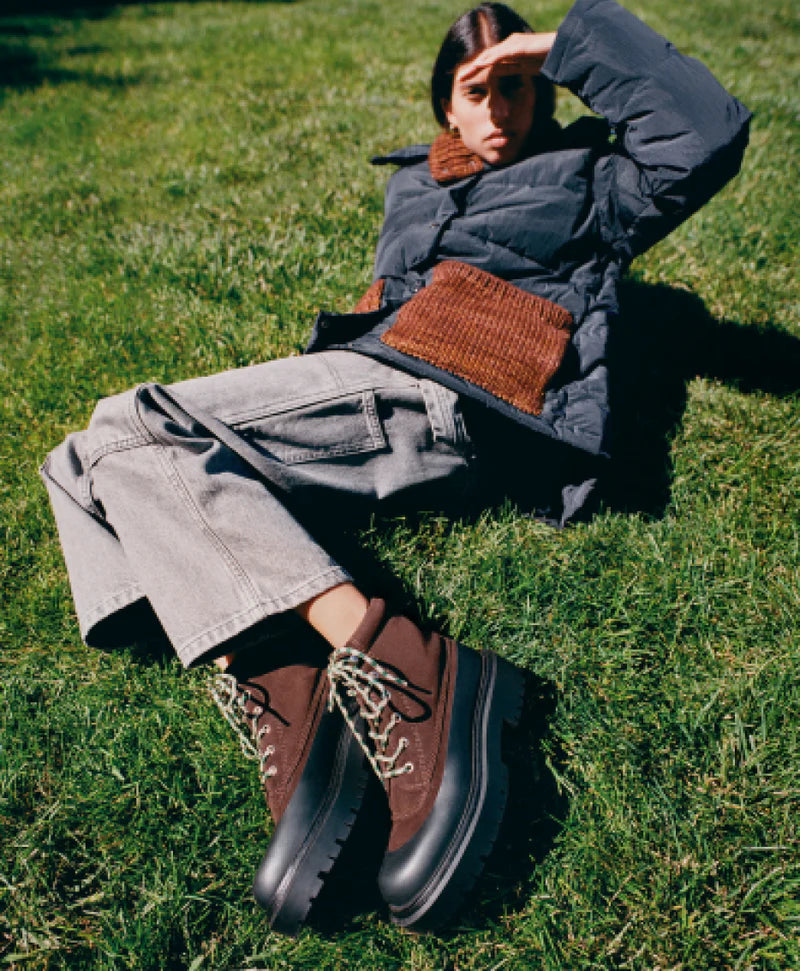 Person sitting on grass wearing a dark coat, gray pants, and brown boots.