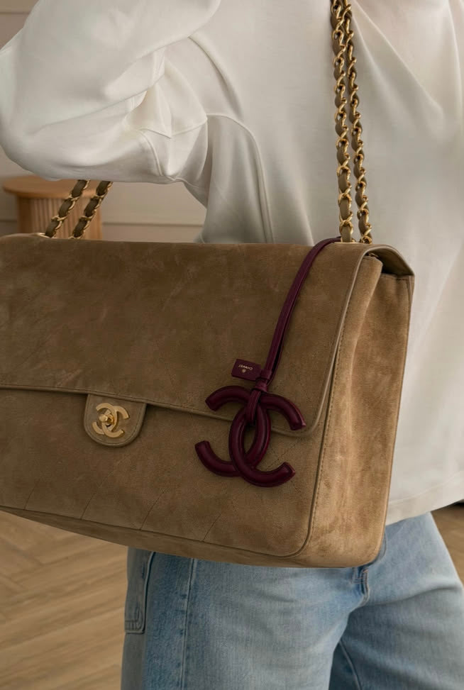 A person wearing a white blouse and light blue jeans carries a taupe suede Chanel shoulder bag with a gold chain strap and a dark burgundy CC turn-lock closure.
