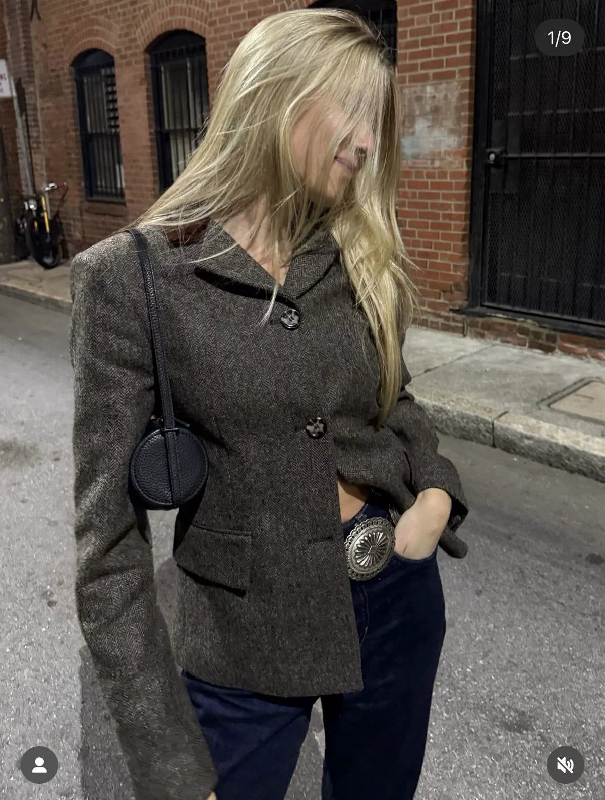 A woman wearing a dark brown textured tweed blazer with dark navy pants, standing on a city street next to a brick building.