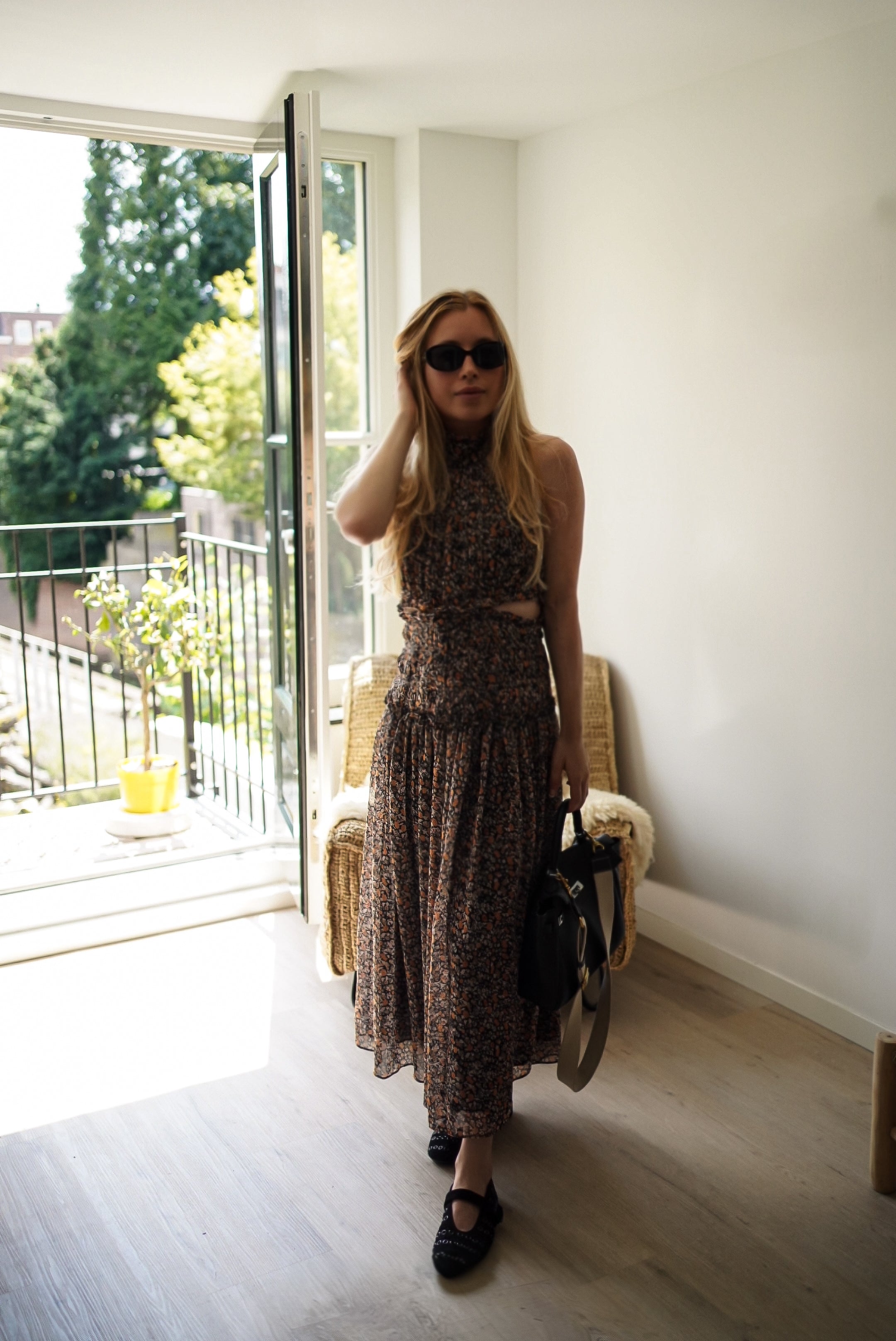 A woman stands in a bright room wearing a sleeveless brown patterned midi dress, black sunglasses, and black Mary Jane shoes, holding a black handbag.